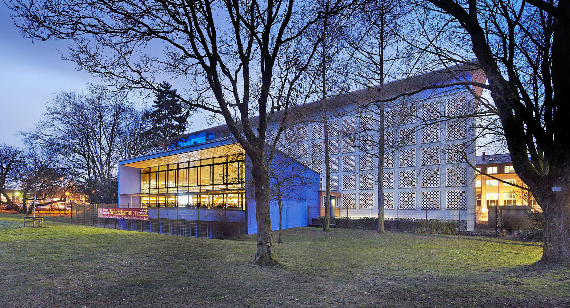 Außenansicht der Schatzkammer der Stadtbibliothek Trier mit modernem Anbau und historischem Bibliotheksgebäude in der Abenddämmerung.